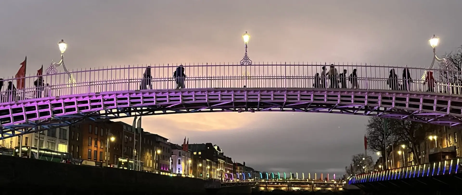 Ha'penny Bridge in Dublin illuminated with purple lights at dusk with pedestrians walking across