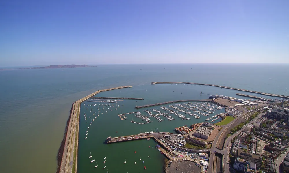 Aerial view of Dun Laoghaire Harbor showing two long piers enclosing numerous boats and yachts, with surrounding buildings and infrastructure under a clear blue sky