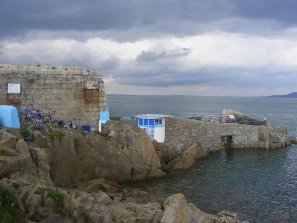 Stone pier at Forty Foot with a small blue and white building, rocky shoreline, parked bicycles, and people near the water under an overcast sky