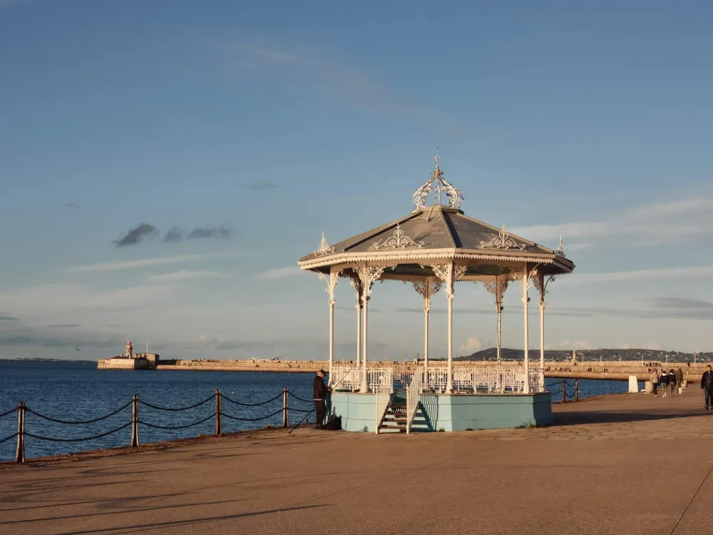 Seaside promenade with a decorative white gazebo, chain-linked fence, lighthouse in the distance, and hills under a clear sky