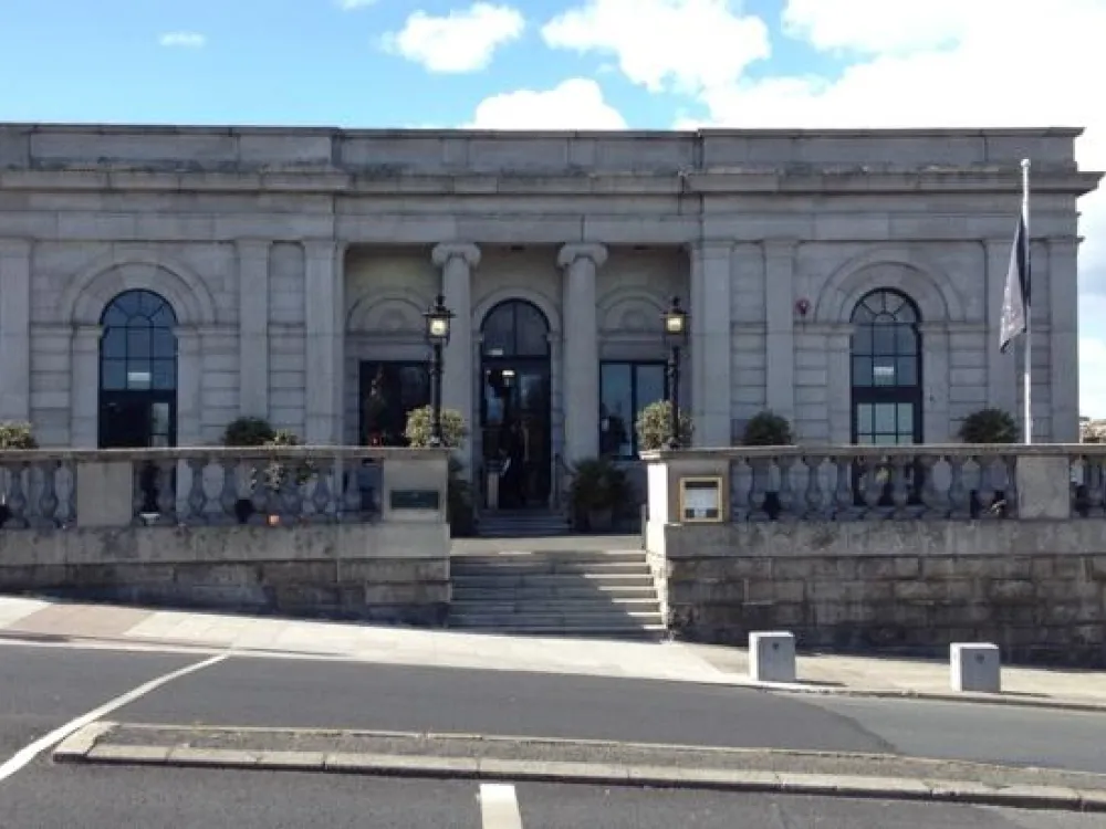 ront facade of a classical stone building with arched windows, columns, stairs, and lantern-style lights under a partly cloudy sky