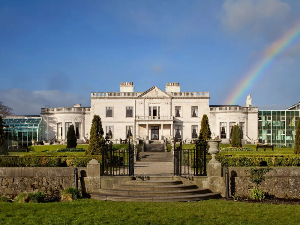 Radisson Blu hotel with classical white facade, manicured garden, stone staircase, and a rainbow arching over a clear blue sky