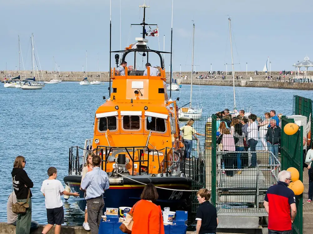 Bright orange lifeboat docked at a pier with people gathered around it in a marina setting